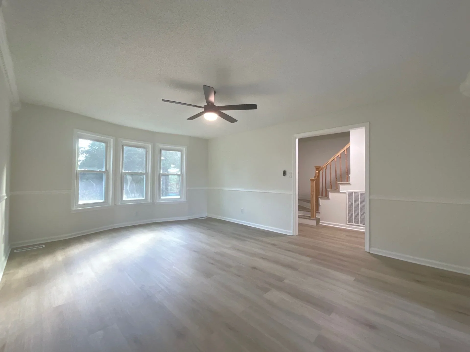 Bright open room with hardwood flooring, white drywall, triple windows, and ceiling fan